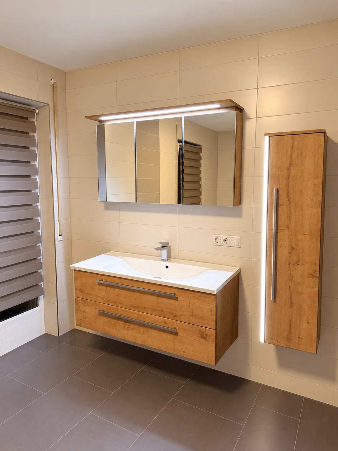 Badezimmer mit hellen Wandfliesen und dunklem Fliesenboden, wandhängender Waschtisch mit Holzunterschrank, beleuchteter Spiegelschrank und Hochschrank in Holzoptik