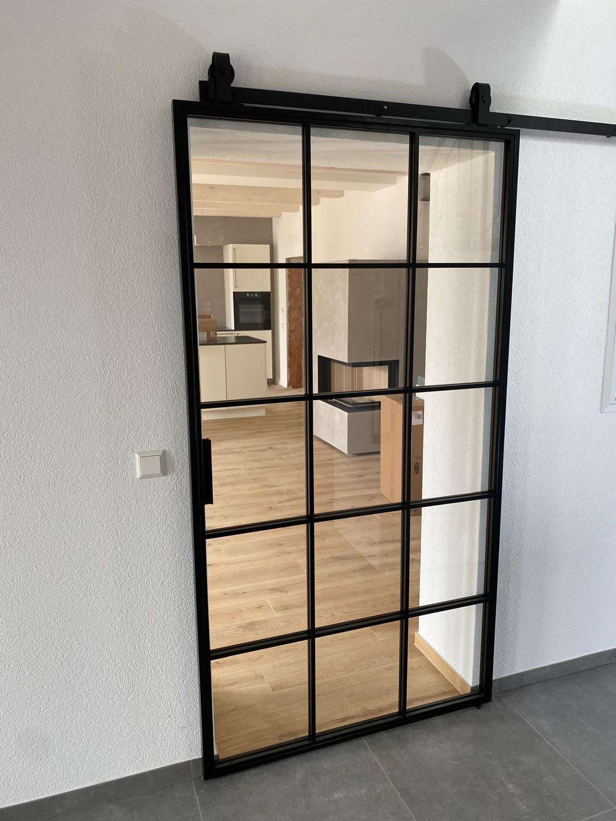 Glas-Schiebetür mit schwarzem Sprossenrahmen an einer offenen Laufschiene, dahinter Küche, Eckkamin und Fliesenboden in Holzoptik