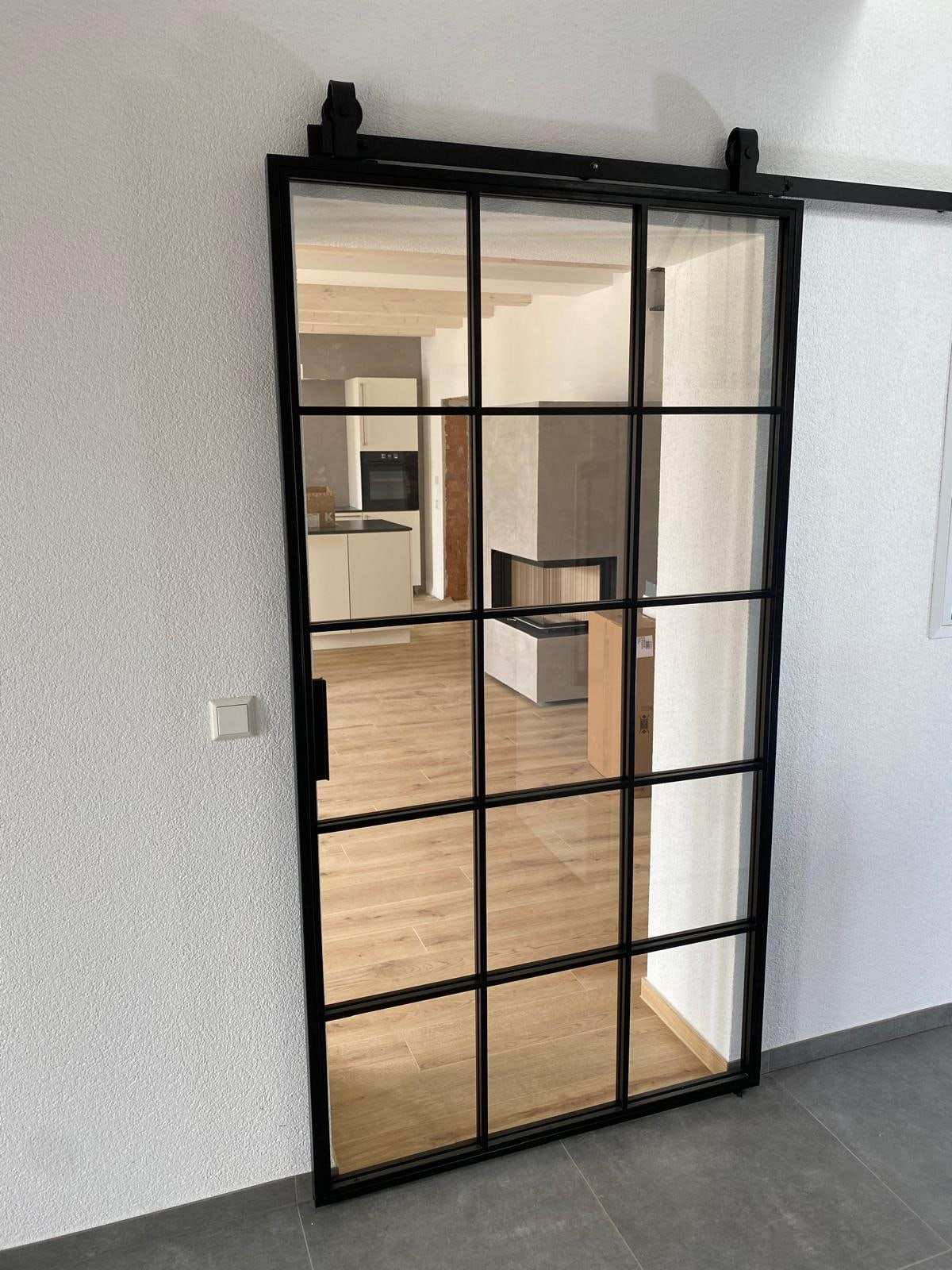 Glas-Schiebetür mit schwarzem Sprossenrahmen an einer offenen Laufschiene, dahinter Küche, Eckkamin und Fliesenboden in Holzoptik