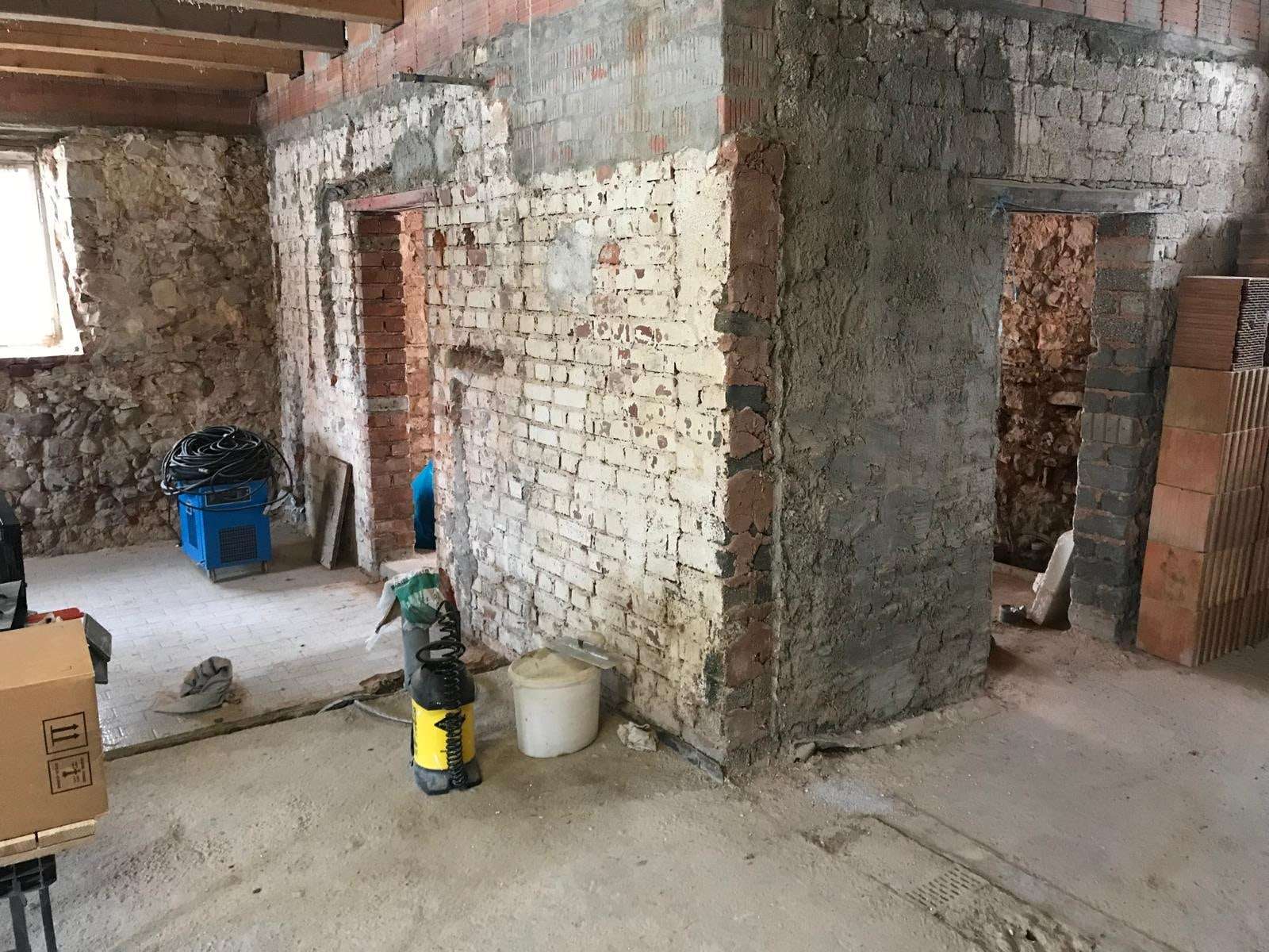 Altbau im Rohzustand: freigelegtes Ziegel- und Natursteinmauerwerk, zwei Türöffnungen mit Stürzen, rechts gestapelte neue Ziegel, Eimer und Werkzeug am Boden